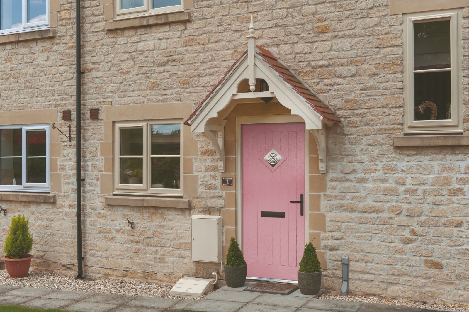 Pink composite front door with diamond glazed panel set within a traditional stone porch of a Cotswold-style cottage, fitted alongside cream uPVC casement windows